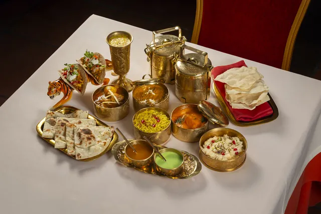 El Lunch box servido en la mesa: fiambreras y cuencos de latón con curry, arroz amarillo, naan, papadum, salsas, kheer y dos Tiger tacos sobre un tigre de cerámica