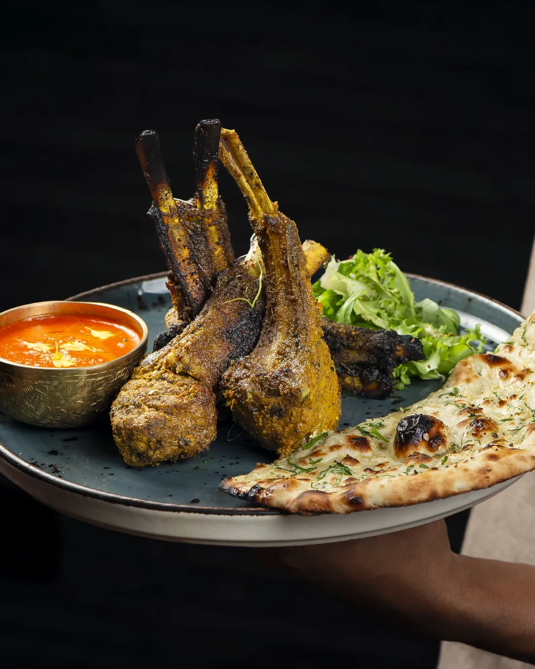 Un plato con chuletas de cordero, naan y salsa, servido en la mano sobre fondo oscuro