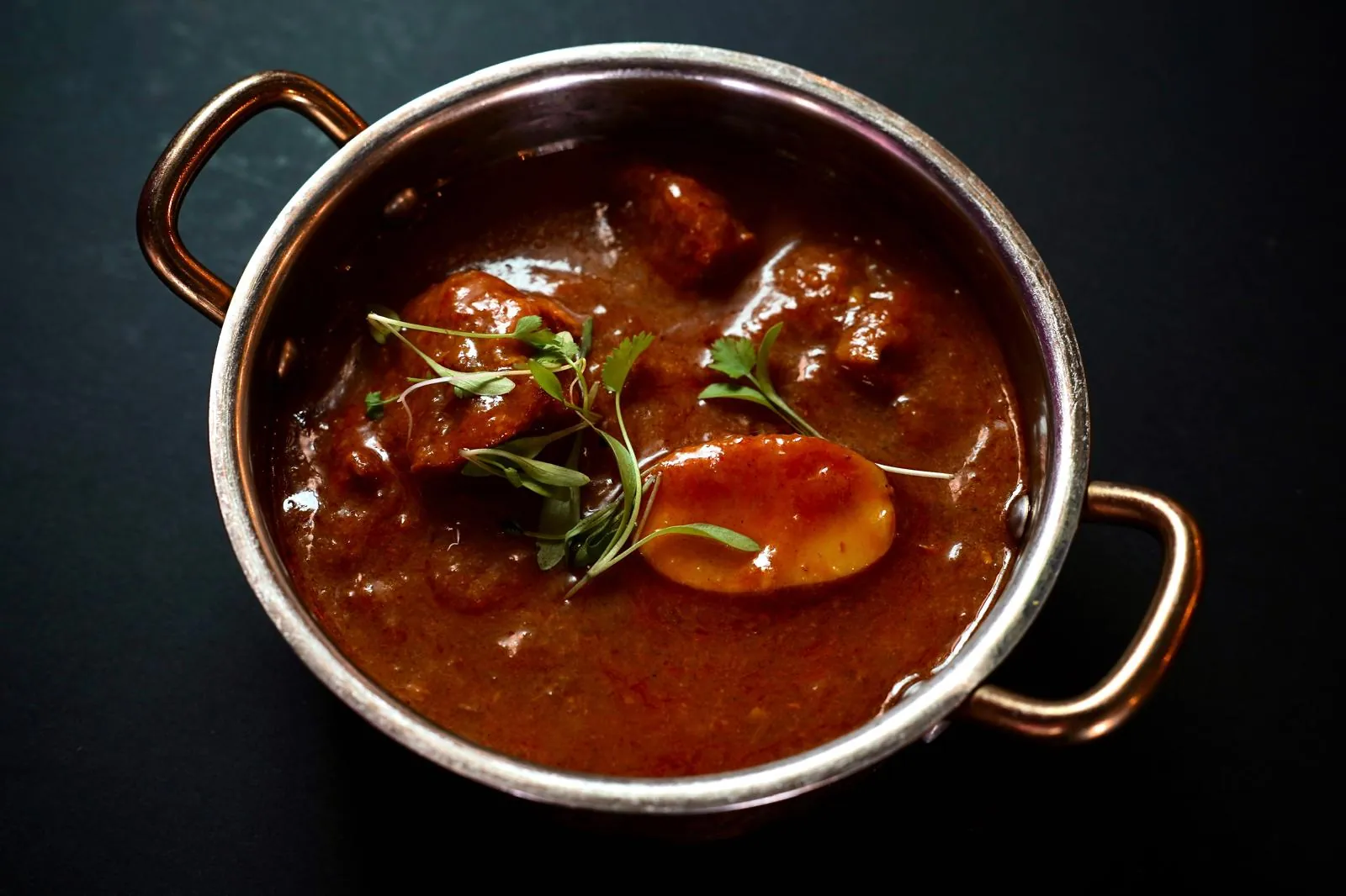 Railway lamb curry en un cazo de cobre visto desde arriba: trozos de cordero y una patata en salsa oscura, con brotes de cilantro