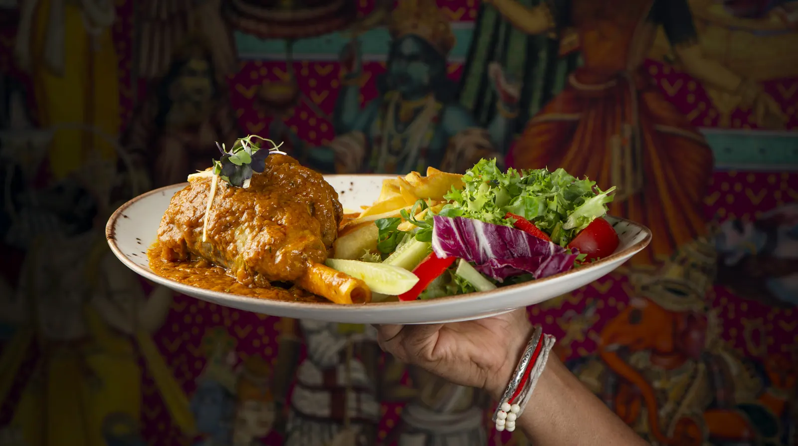 Un plato de cordero en salsa con ensalada, sostenido en alto delante de un mural indio de colores